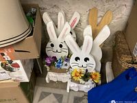 Front view of multiple white wooden Easter bunnies with pink ears and flower garlands, standing upright for outdoor decoration.