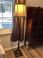 Full view of floor lamp turned on next to window and wooden cabinet.