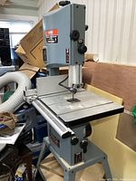 Front-angle view showing bandsaw on steel stand with sliding rip fence and dust collection hose