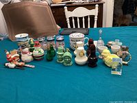 Photo showing assorted knickknacks arranged in rows including various salt and pepper shakers, small cups, Christmas figures, glass shot glasses, and miscellaneous decorative objects on a table with blue green tablecloth and chairs in the background.