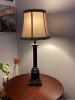 Photo of a black table lamp with a beige fabric shade trimmed in black, placed on a round wooden table against a light gray wall.