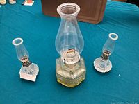 Three vintage oil lamps displayed on a blue cloth surface with a brown background. Center lamp is bigger with a wide base, flanked by two smaller lamps with ribbed glass chimneys.