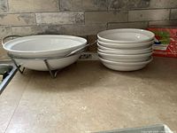 White ceramic pasta serving dish on silver metal stand with six white pasta bowls stacked beside it on countertop.
