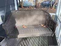 Front view of the brown resin wicker porch swing showing seat and backrest with a few dry leaves on the surface.