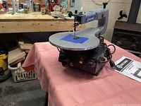 Front angle view of King Canada 16-inch scroll saw on a pink cloth-covered table with surrounding workshop items.