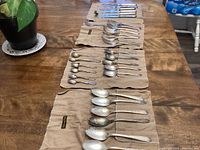 Top view photo showing multiple rows of silver-plated cutlery including spoons and forks laid out on cloth holders on wooden table with a potted plant nearby.