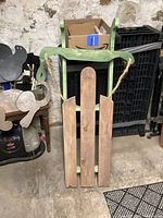 Wooden sleigh leaning against a stone wall and surrounded by other items in a basement or storage area, showing front view with rope handles and three wooden slats.