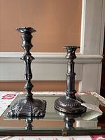 Two silver plate candlesticks displayed side by side on a mirrored surface, showing height and ornate base details.