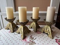 Four gilded bronze tripod candleholders with ivory pillar candles on fabric surface with floral embroidery.