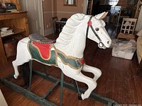 Side view of white painted carousel hobby horse with green and red painted saddle decorations, leather saddle, horsehair tail on dark wood base frame with rocker support