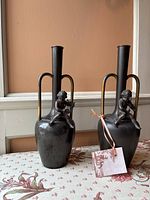 Two dark patinated bronze urns each topped with a seated cherub and featuring tall tubular necks with arched handles.
