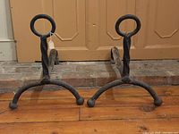 Pair of antique iron andirons side by side on wooden floor in front of brick hearth and wooden panel wall.
