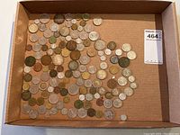 Wide shot showing a large number of assorted foreign coins of varying sizes and colors in a cardboard box.