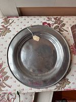 Full view of large peened pewter serving platter with beaded edge on floral fabric surface showing metallic sheen and hammered texture.