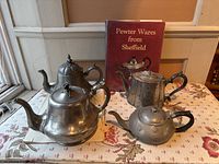 Four pewter teapots displayed with a red reference manual 'Pewter Wares from Sheffield' on a patterned surface near a window.