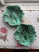 Two green cabbage leaf-shaped ceramic plates on floral fabric surface, top view showing detailed texture and veining.