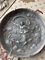 Front view of the round pewter plaque with raised winged woman and celestial design, placed on a patterned fabric surface.