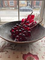 Pewter bowl with copper rim holding decorative glass grapes with red leaves, photographed by window with outdoor background visible.