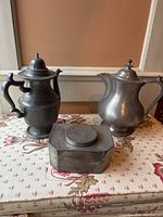 Photo showing two large pewter coffee pots and one decorative pewter box on floral fabric surface.