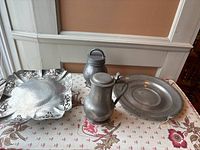 Four antique pewter pieces arranged on a fabric surface: hammered tray, jug, container and plate.