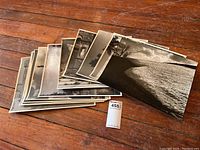 Group image showing a stack of vintage black and white photographic prints with a MaxSold tag nearby, displaying wear and varied glossy/matte surfaces.