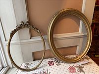 Two gold oval wooden frames side by side on a floral cloth. One frame has ornate bow and floral decoration at top, the other is simpler with beaded edge detail.