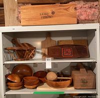 Multiple wooden kitchen items on a shelving unit including small bowls, large bowl, serving platter, cutting boards, cheese board, shoe shine kit, fruit bowl, maracas, and a wooden crate.