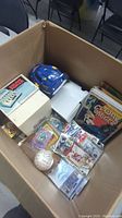 View of collectibles in a cardboard box showing a signed baseball, sports cards, comic books and magazines.