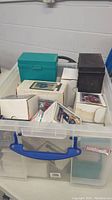 Large clear plastic bin filled with various white cardboard boxes, a green card storage box, and a black card holder containing numerous sports cards.