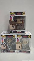 Three Funko Pop boxes stacked: Nancy on top, Eddie and Henry underneath, all from Stranger Things.