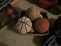Photo showing 6 used basketballs in a cardboard box on a floor, various colors and brands including one Spalding ball.