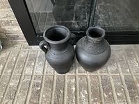 Two black decorative vases placed on a hearth in front of a fireplace. Both are matte black with different shapes and surface textures.
