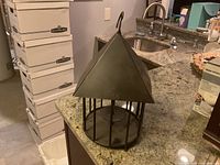 Black metal candle cage lantern with triangular roof placed on kitchen counter, visible vertical bars, circular base, and hanging hook at the top.