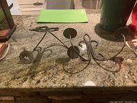 Pair of black iron wall-mount candle holders displayed on a granite countertop, showing curved arms and flat circular bases for holding candles.