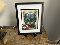 Framed picture of a cat wearing a Captain America themed mask and shield, displayed on a stand on a table.