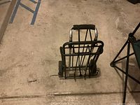 Black folding cart with metal frame and two wheels, viewed folded and standing upright on a concrete floor.