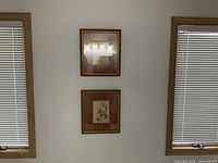 Two decorative framed flower pictures hanging on a wall between two windows with blinds.