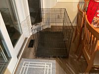 Black metal wire dog crate placed indoors on a wooden floor near glass doors and dining furniture.