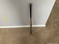 Full view of an Easton baseball bat standing upright against a wall showing gray and orange barrel with black grip handle on carpet flooring.