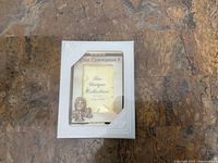 Small white picture frame with religious designs on the corner indicating it is for a First Communion photograph. The frame is in clear packaging showing it is unused.