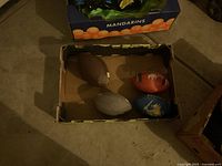 Photo of four used footballs in a cardboard box on a floor, including brown, gray, red, and blue footballs.