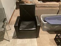 Front view of black faux leather gaming chair with cushioned seat and armrests in a room with tiled floor and nearby plastic bins.