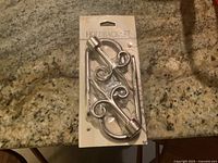 Photo of two silver-tone metal curtain holdbacks with included mounting screws in original packaging, placed on granite countertop.