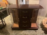 Photo of a dark wooden jewelry box with two glass-paneled doors on either side and four drawers in the center with metal handles, placed on a table.
