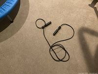 Photo of black jump rope with plastic ergonomic handles lying on carpet next to a blue trampoline.