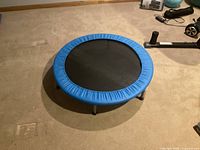 Photo of a small round mini trampoline with a blue padded frame cover and six metal legs on a carpeted floor, with some other fitness items visible in background.
