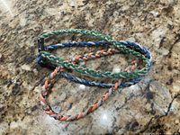 Three braided rope necklaces in green, blue, and orange with black plastic clasps, laid on a stone countertop.