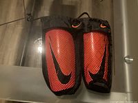 Photo of a pair of red Nike soccer shin guards with black Nike swoosh logos, including black mesh sleeves with orange Nike swoosh logo for securing them.