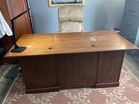Front view of a solid wood desk with a light beige office chair behind it, positioned on a patterned rug.