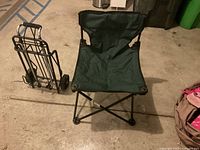 Green folding sports chair set on concrete floor with foldable black metal frame and green fabric seat. A hand truck is nearby.